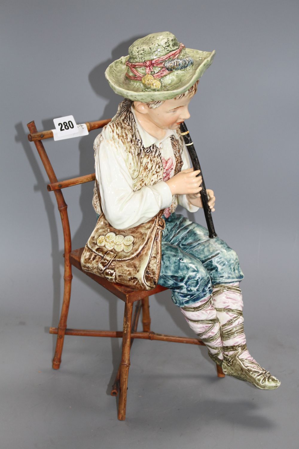 A late 19th century Austrian tin glaze pottery figure of a Tyrolean boy playing a flute, seated upon a bamboo wood chair, height 43cm